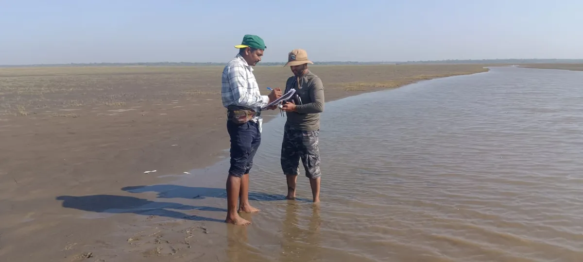 Two field team members standing at the edge of a shallow tidal channel on a wide mudflat, one writing on a clipboard.